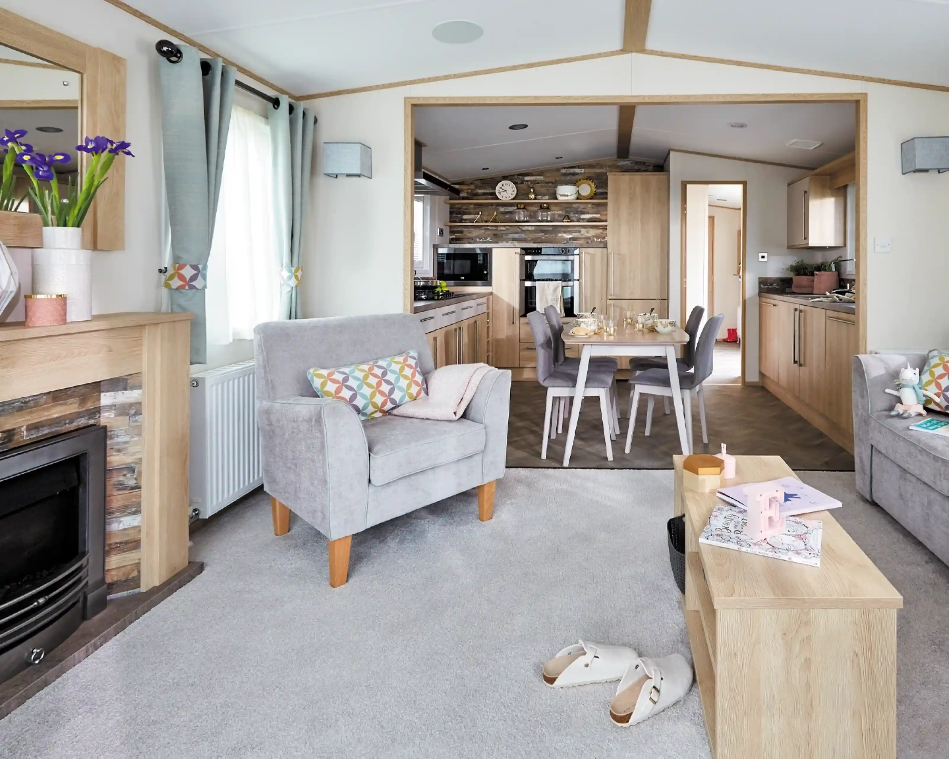A cozy living room features two gray armchairs with colorful cushions, a wooden coffee table, and a modern electric fireplace. In the background, a well-equipped kitchen is visible, with wooden cabinetry and a dining area set for a meal. Light filters through large windows, adding a warm ambiance to the space.