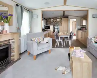 A cozy living room features two gray armchairs with colorful cushions, a wooden coffee table, and a modern electric fireplace. In the background, a well-equipped kitchen is visible, with wooden cabinetry and a dining area set for a meal. Light filters through large windows, adding a warm ambiance to the space.