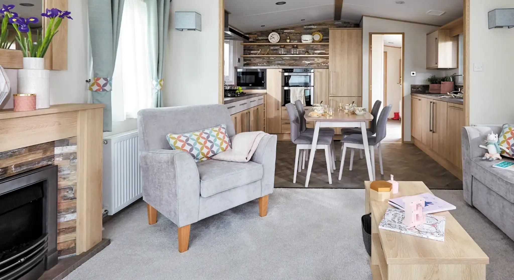 A cozy living room features two gray armchairs with colorful cushions, a wooden coffee table, and a modern electric fireplace. In the background, a well-equipped kitchen is visible, with wooden cabinetry and a dining area set for a meal. Light filters through large windows, adding a warm ambiance to the space.