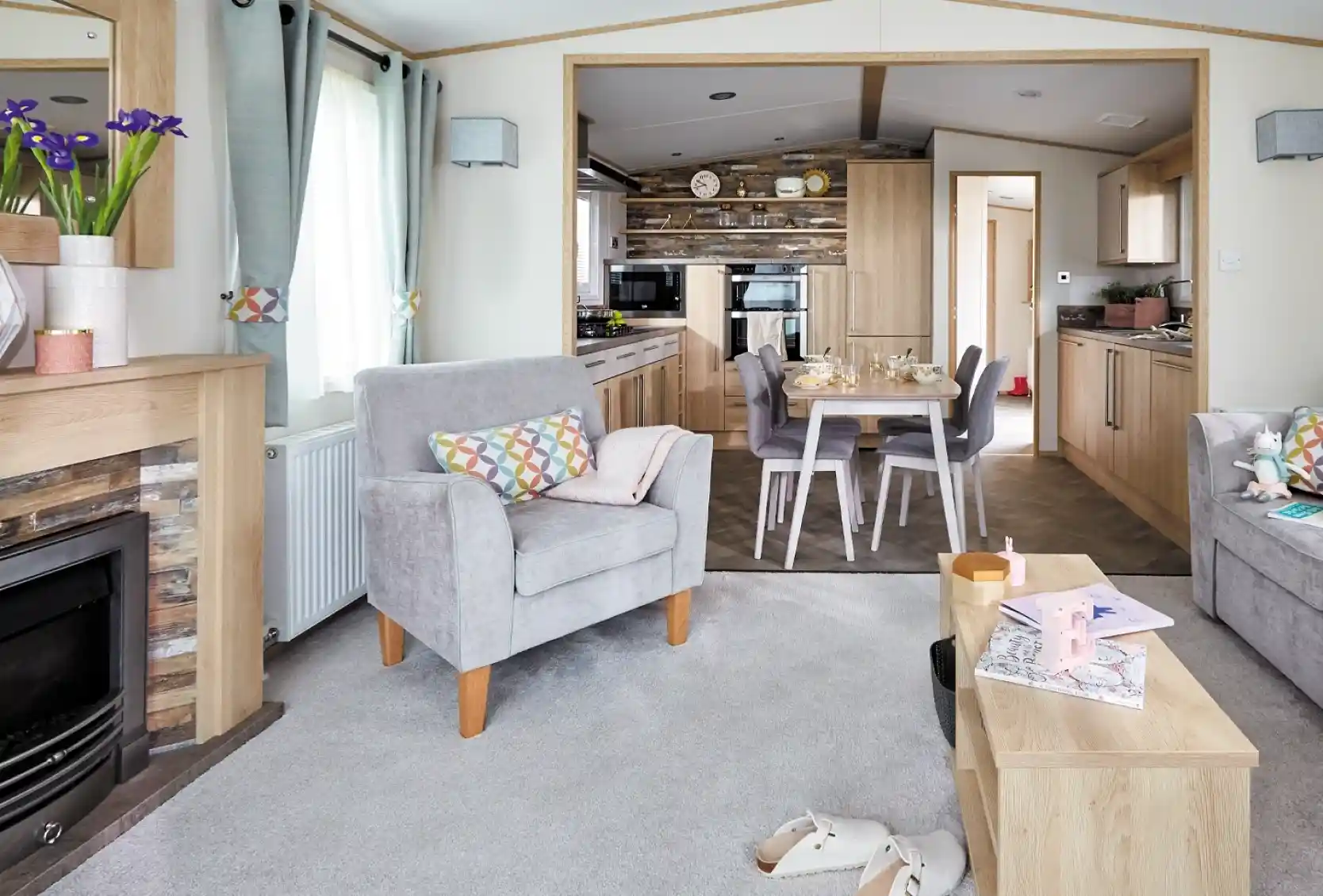 A cozy living room features two gray armchairs with colorful cushions, a wooden coffee table, and a modern electric fireplace. In the background, a well-equipped kitchen is visible, with wooden cabinetry and a dining area set for a meal. Light filters through large windows, adding a warm ambiance to the space.