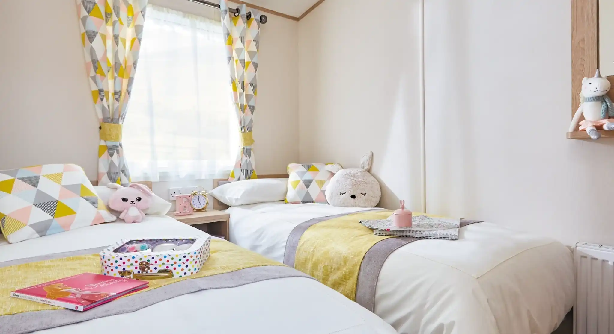 A cozy bedroom featuring two twin beds adorned with white linens and yellow and gray striped accents. Geometric patterned curtains hang at the window, and each bed has decorative pillows and plush toys. A mirror and a small table with books and a colorful box are visible in the room.