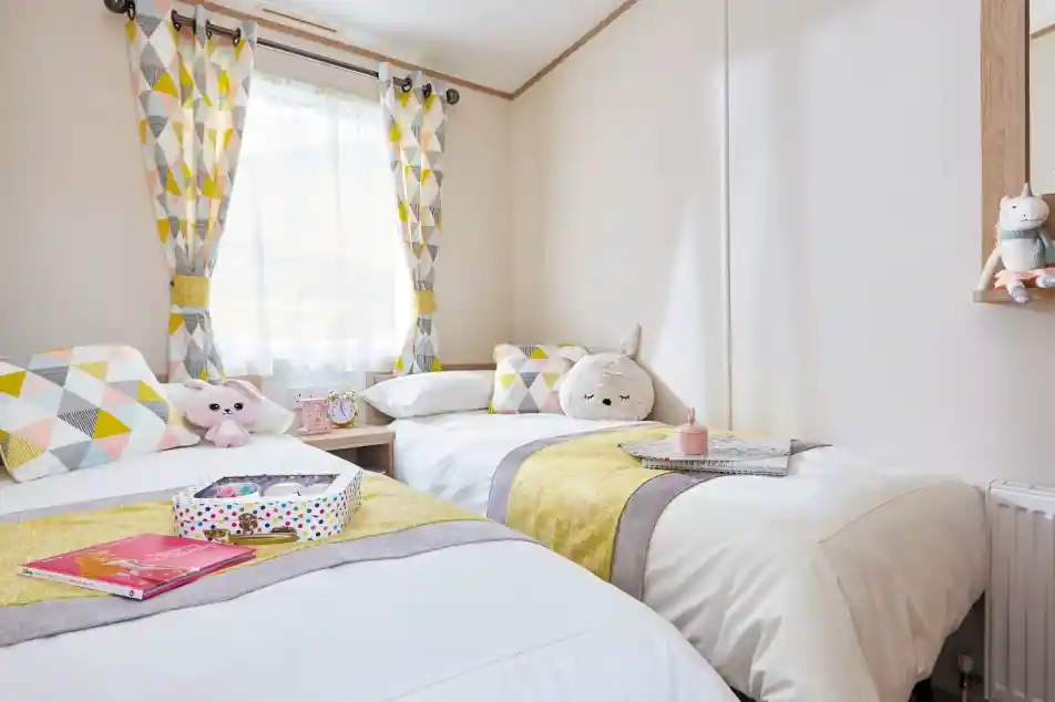A cozy bedroom featuring two twin beds adorned with white linens and yellow and gray striped accents. Geometric patterned curtains hang at the window, and each bed has decorative pillows and plush toys. A mirror and a small table with books and a colorful box are visible in the room.