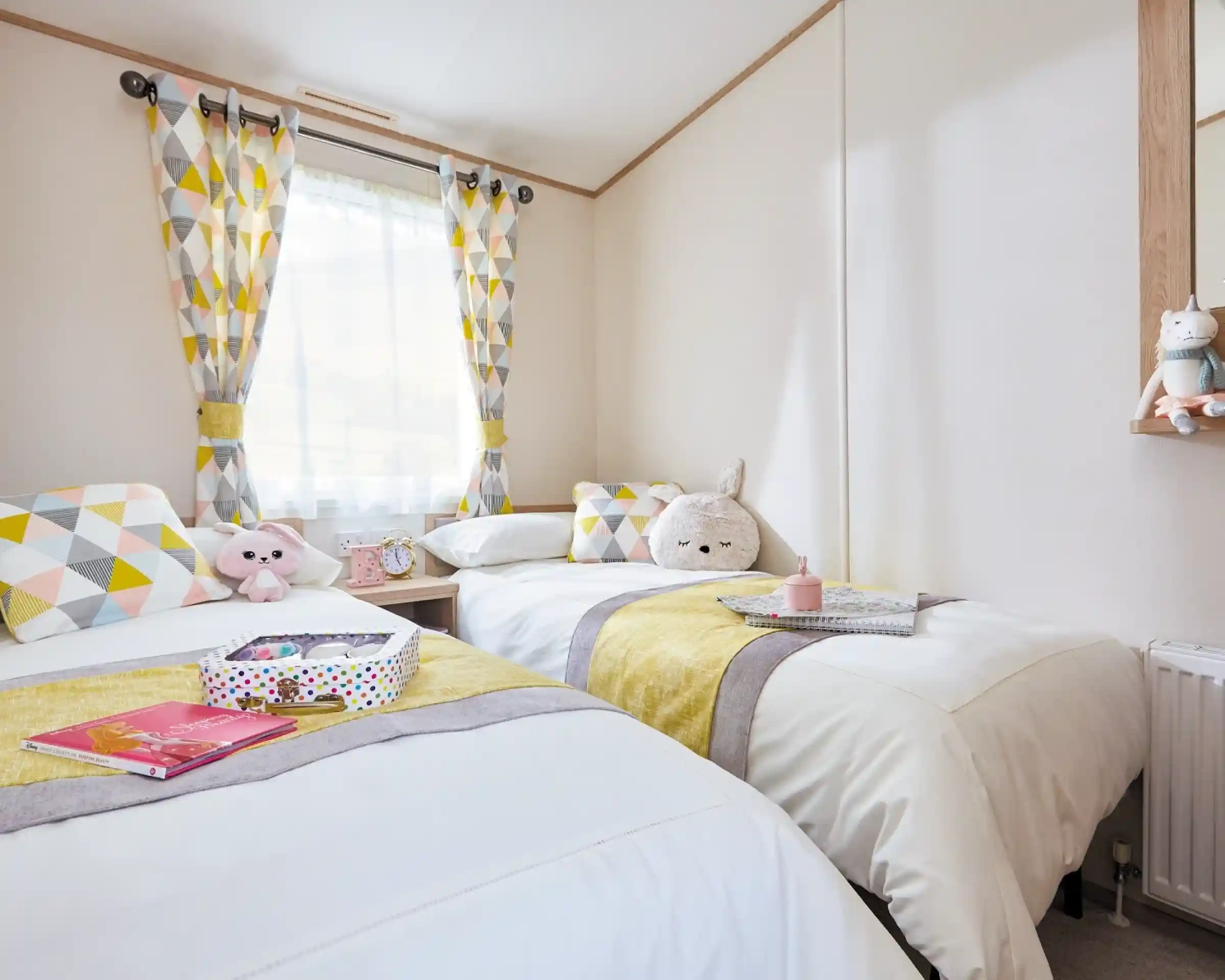 A cozy bedroom featuring two twin beds adorned with white linens and yellow and gray striped accents. Geometric patterned curtains hang at the window, and each bed has decorative pillows and plush toys. A mirror and a small table with books and a colorful box are visible in the room.