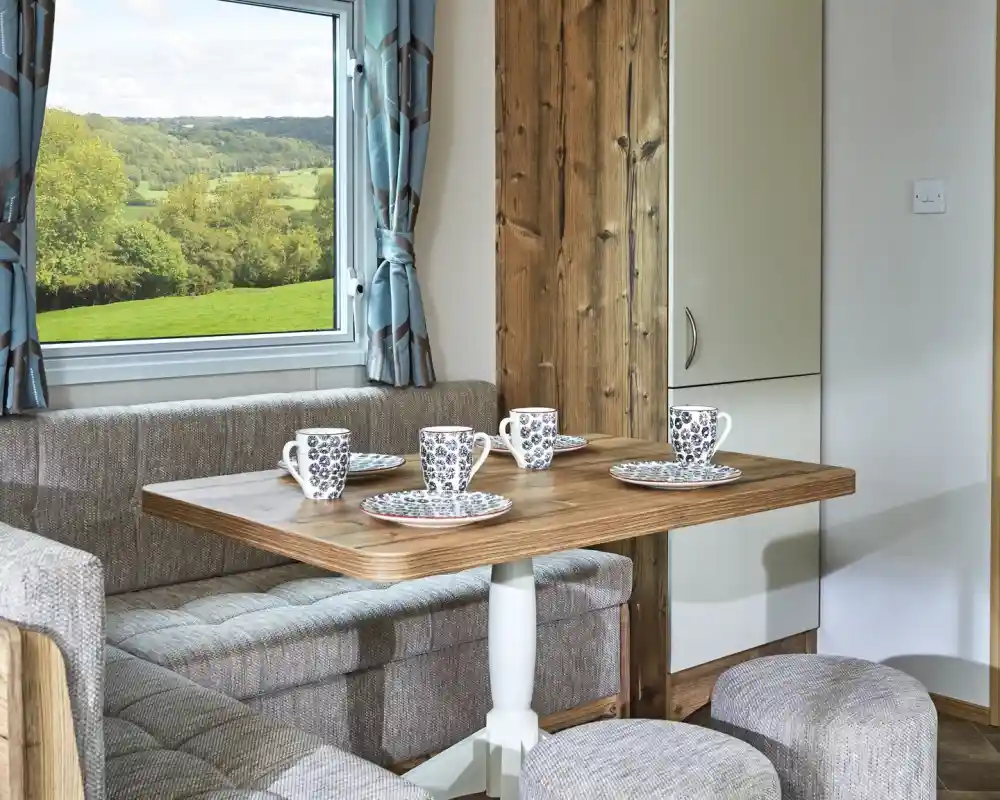 Cosy dining nook with a wooden table, upholstered seating, and mugs set for coffee with a scenic view through the window.