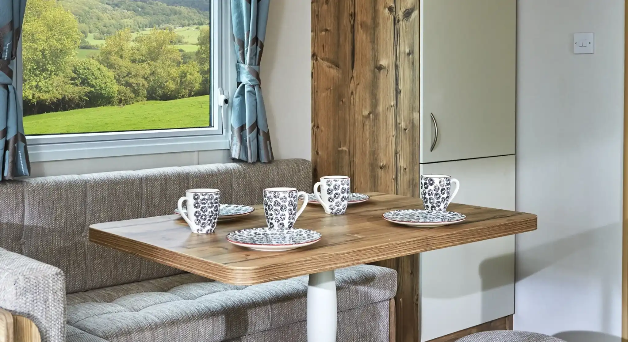 Cosy dining nook with a wooden table, upholstered seating, and mugs set for coffee with a scenic view through the window.