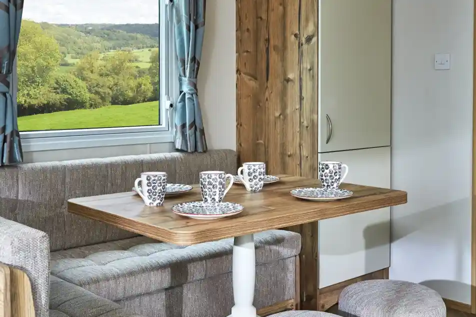 Cosy dining nook with a wooden table, upholstered seating, and mugs set for coffee with a scenic view through the window.