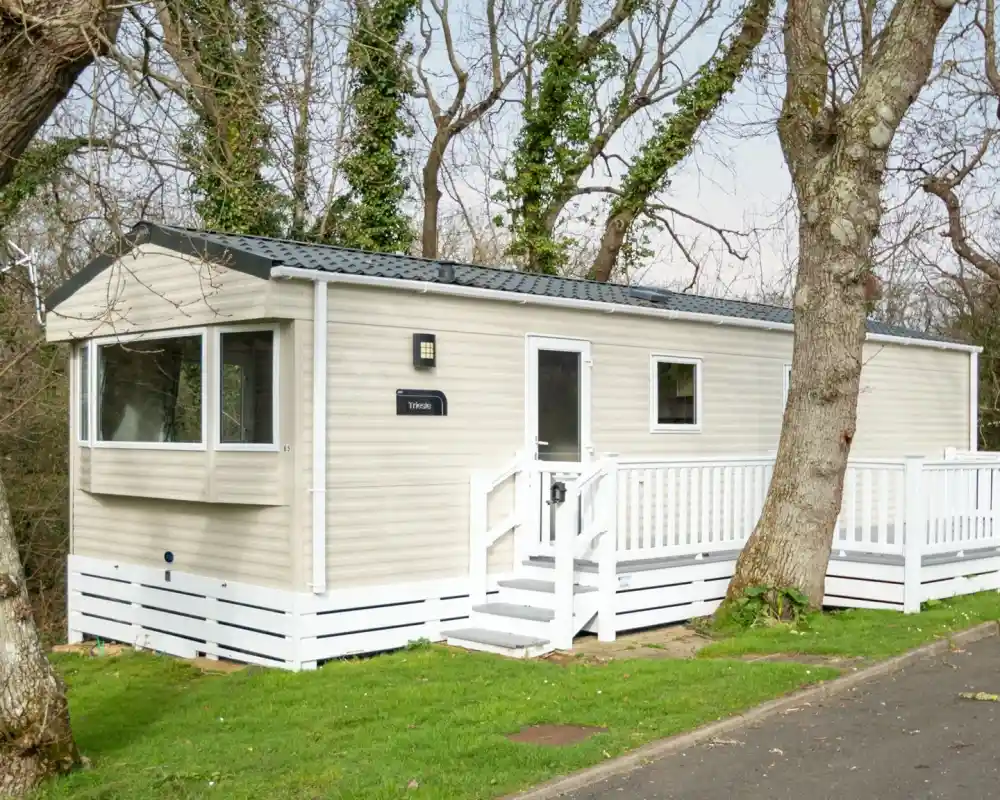 A beige mobile home with a white deck and railing sits amongst trees.