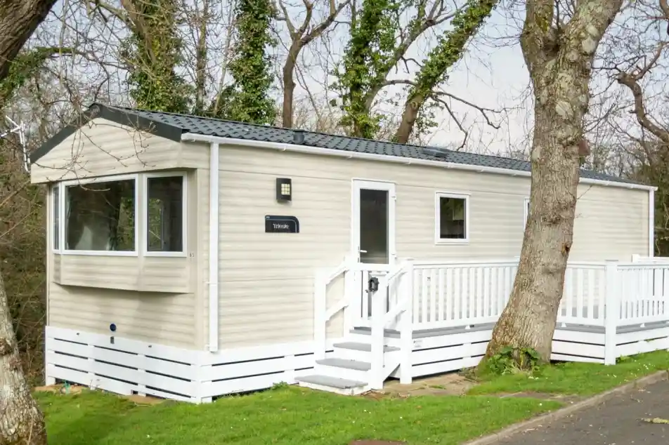 A beige mobile home with a white deck and railing sits amongst trees.