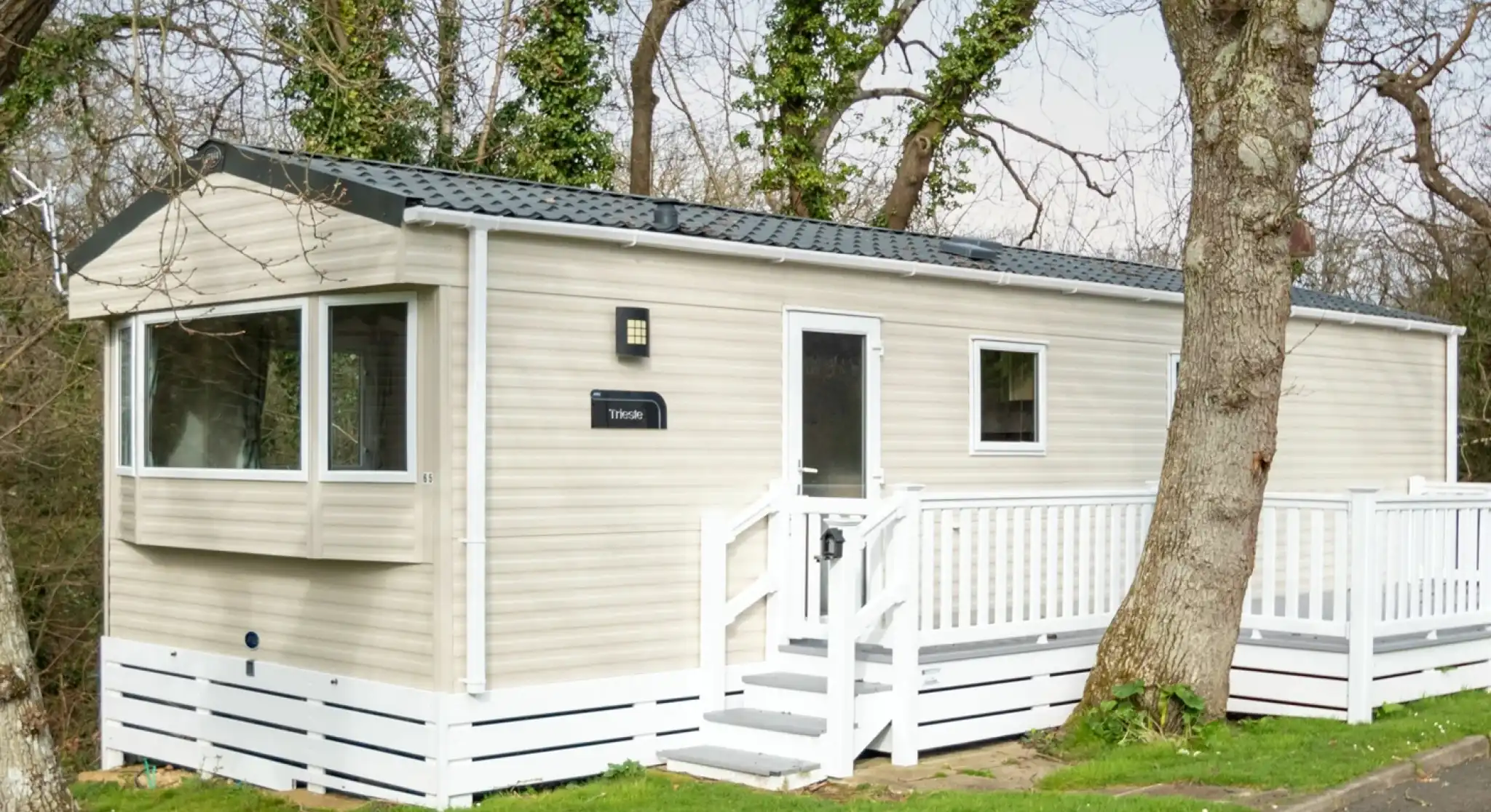 A beige mobile home with a white deck and railing sits amongst trees.