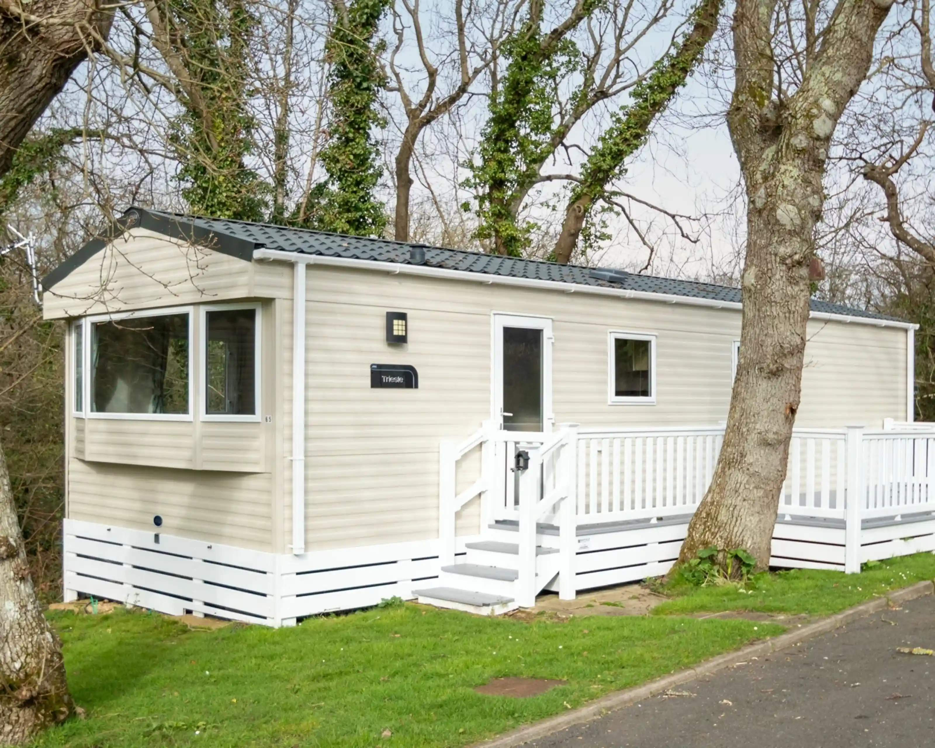A beige mobile home with a white deck and railing sits amongst trees.
