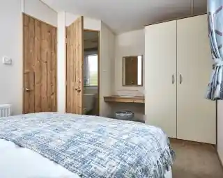 A bedroom with a blue patterned blanket on the bed, wooden doors, and a cream wardrobe with a mirror above a small vanity.