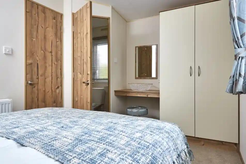 A bedroom with a blue patterned blanket on the bed, wooden doors, and a cream wardrobe with a mirror above a small vanity.