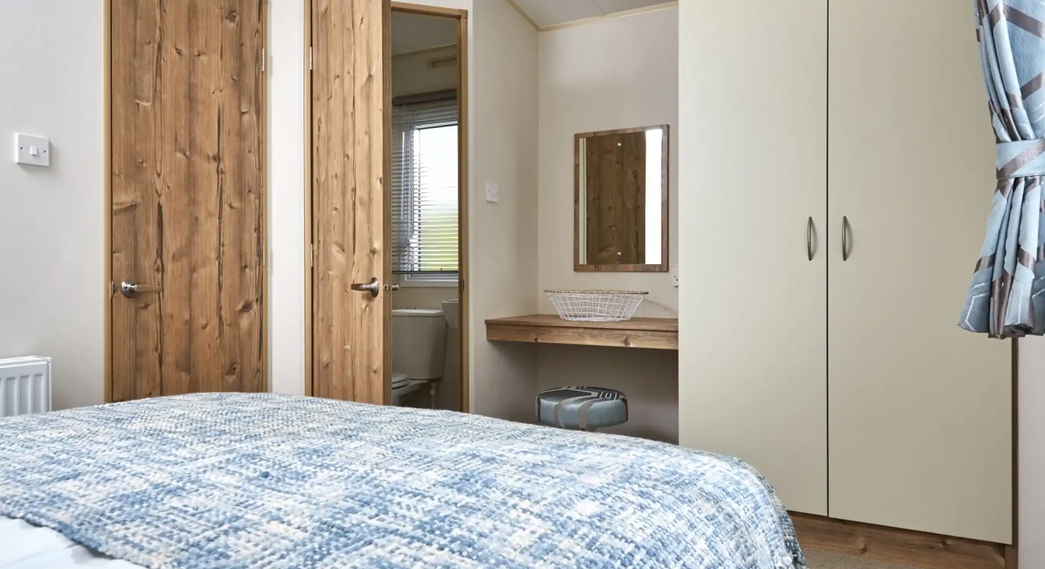 A bedroom with a blue patterned blanket on the bed, wooden doors, and a cream wardrobe with a mirror above a small vanity.