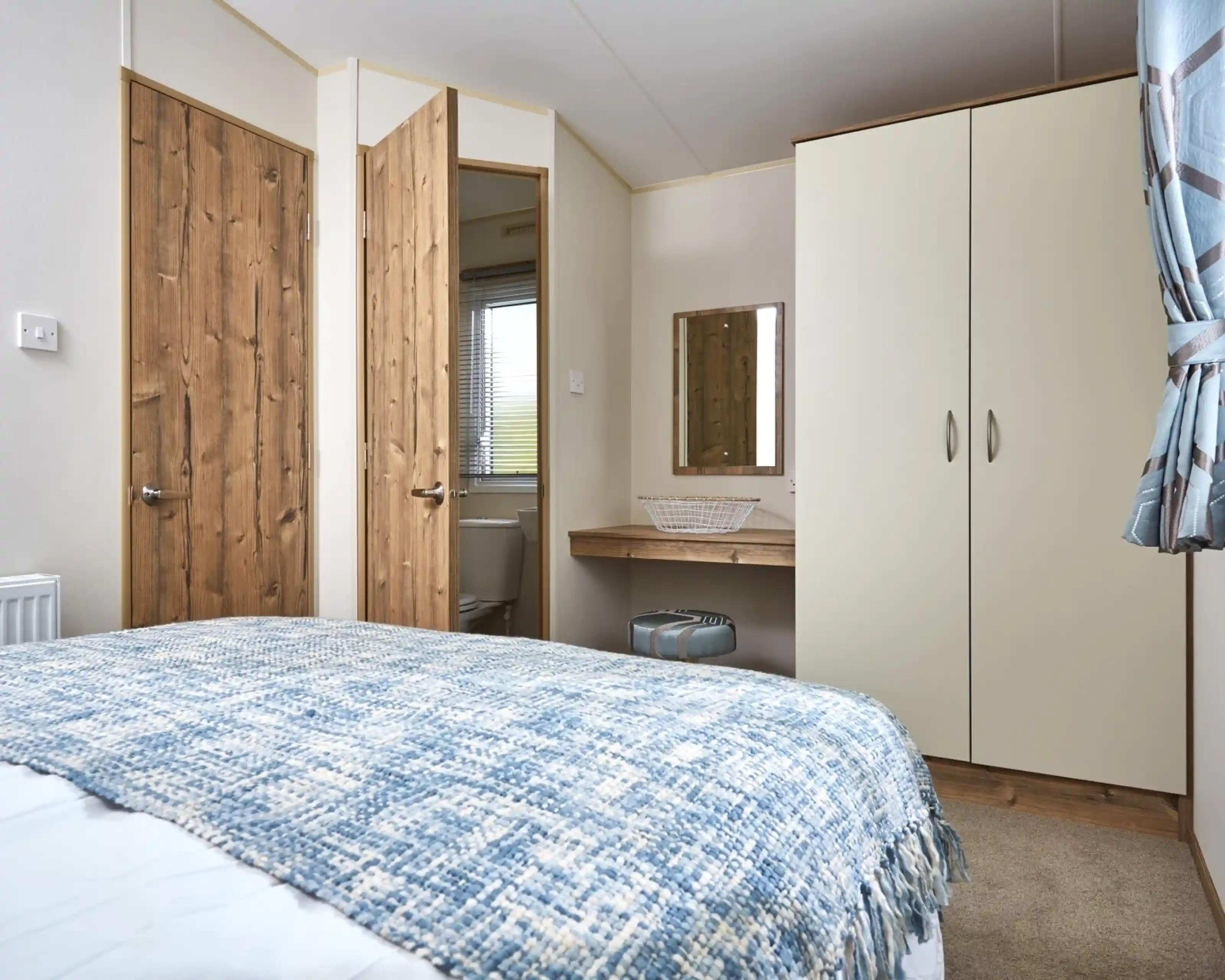 A bedroom with a blue patterned blanket on the bed, wooden doors, and a cream wardrobe with a mirror above a small vanity.