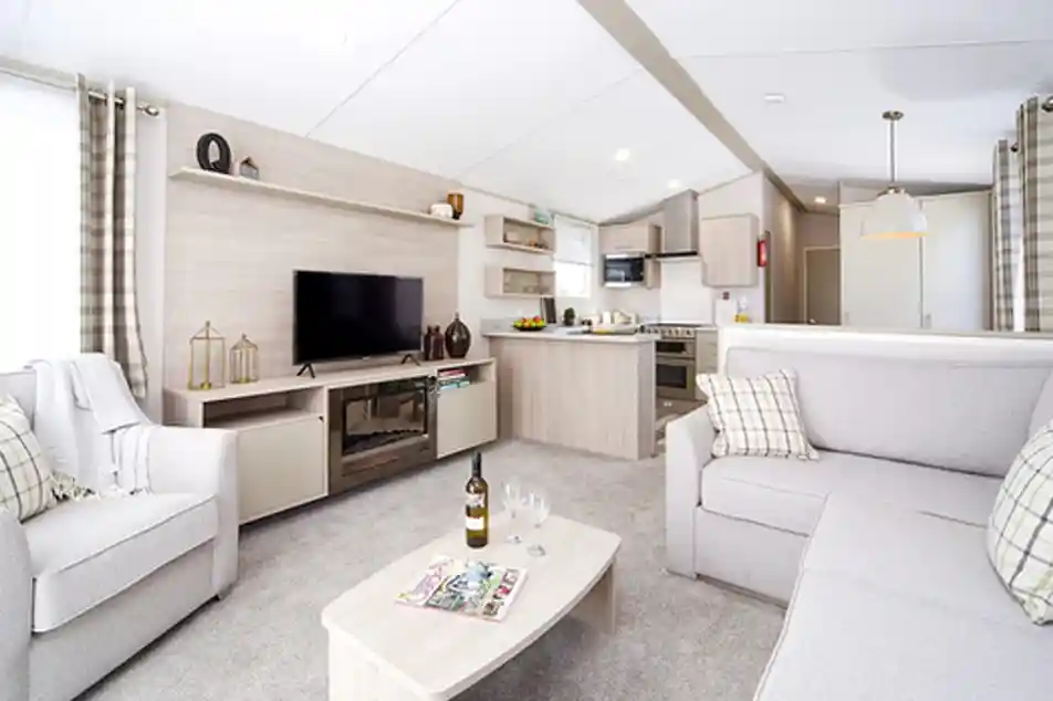 Modern living room and kitchen area with light grey sofas, a TV stand with a black television, and a neutral-toned kitchen with appliances.