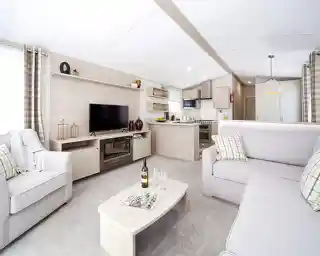 Modern living room and kitchen area with light grey sofas, a TV stand with a black television, and a neutral-toned kitchen with appliances.