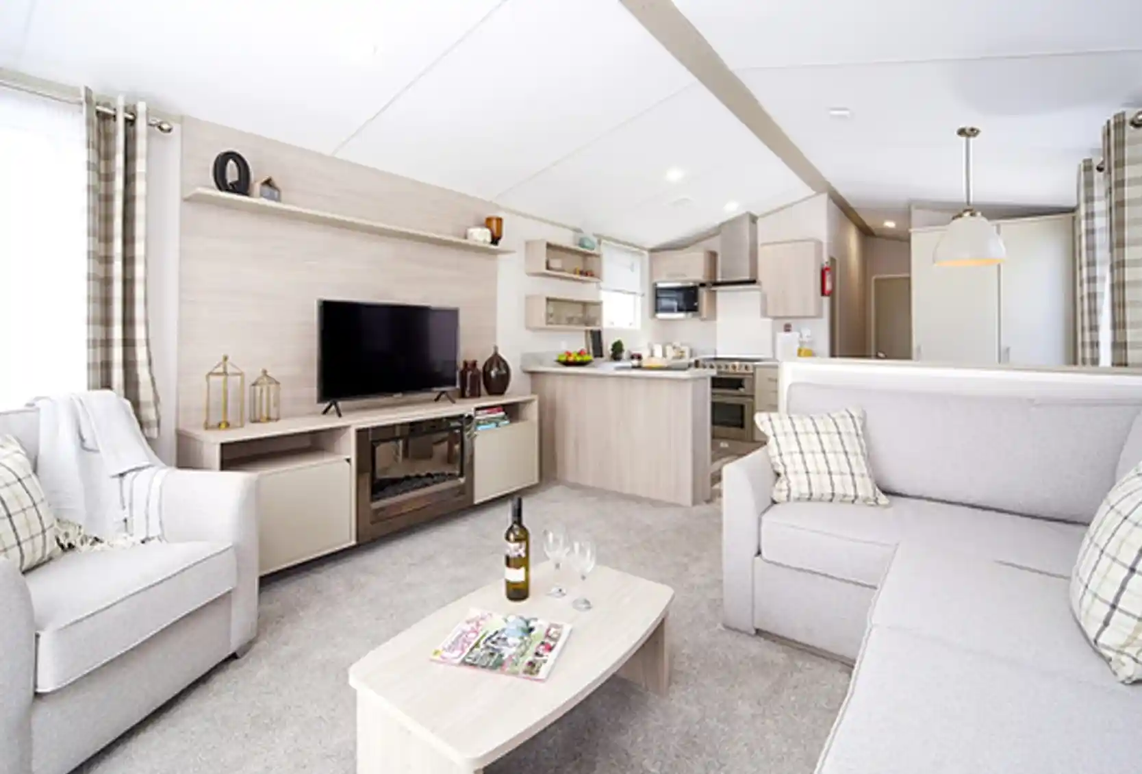 Modern living room and kitchen area with light grey sofas, a TV stand with a black television, and a neutral-toned kitchen with appliances.