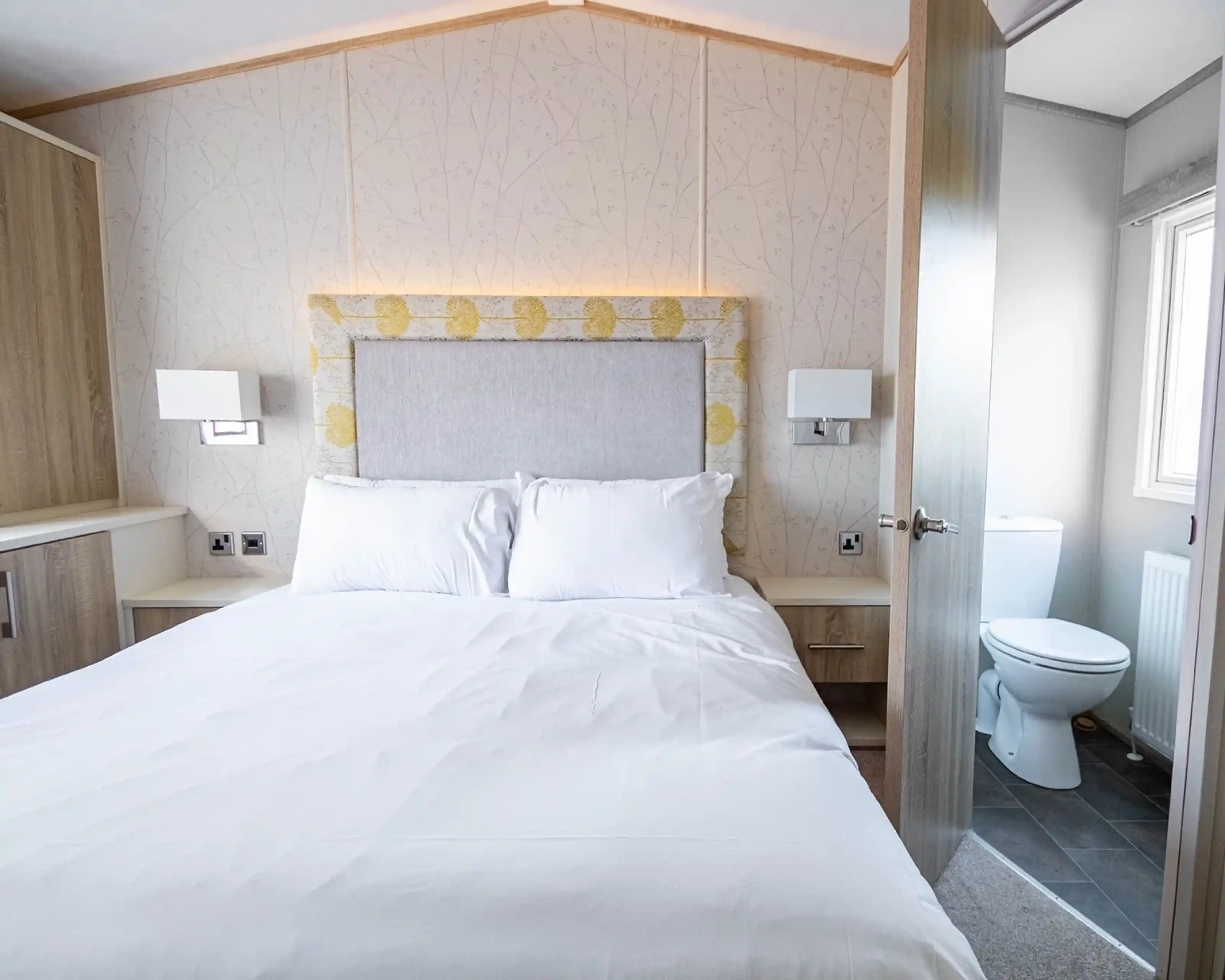 A cozy bedroom featuring a double bed with white linens, a padded headboard, and sleek bedside lamps. A door on the right leads to a small, clean bathroom with a toilet visible. The room has light-colored wooden cabinetry and a soft floral wallpaper.