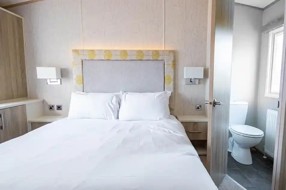 A cozy bedroom featuring a double bed with white linens, a padded headboard, and sleek bedside lamps. A door on the right leads to a small, clean bathroom with a toilet visible. The room has light-colored wooden cabinetry and a soft floral wallpaper.