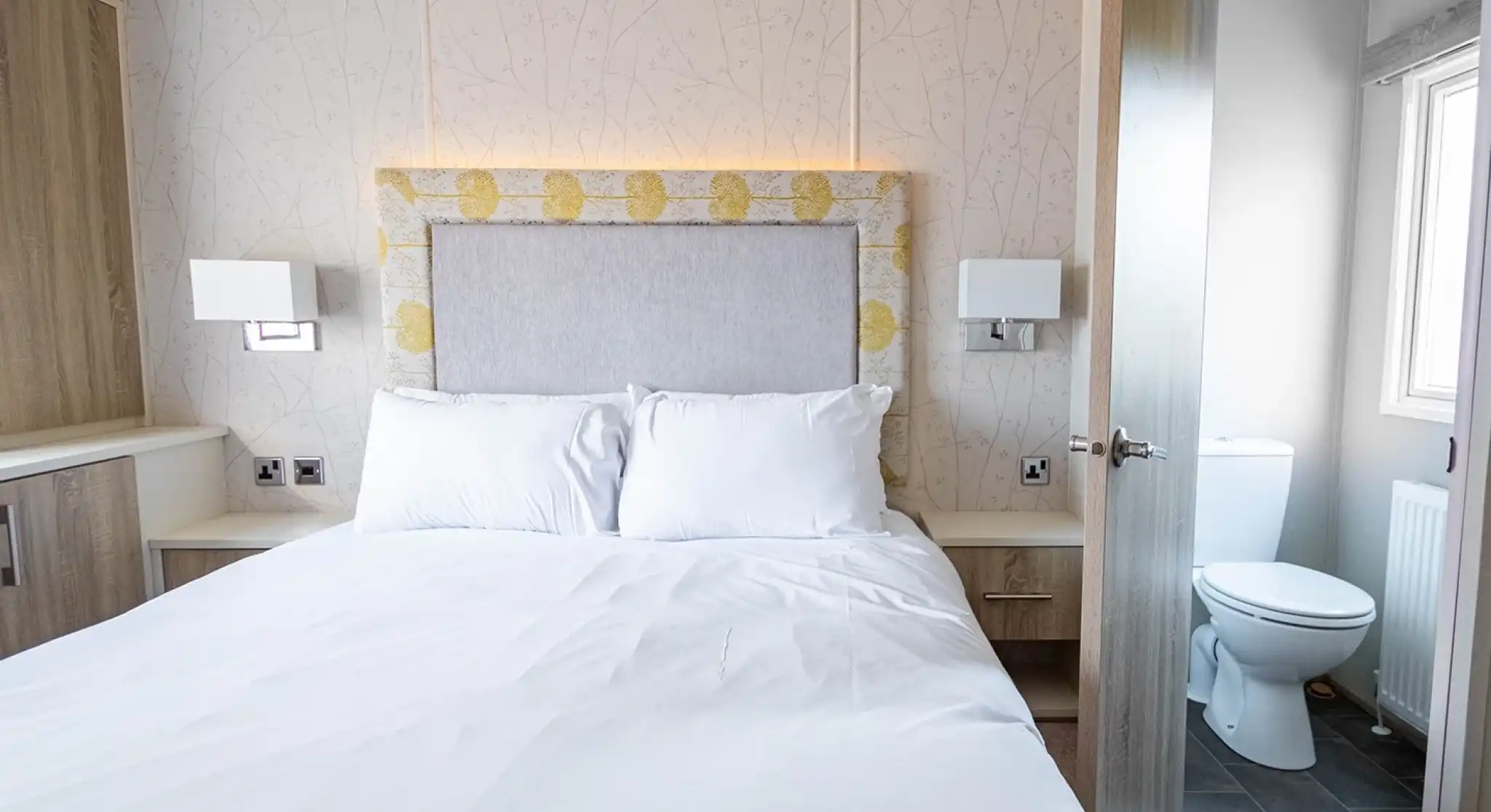 A cozy bedroom featuring a double bed with white linens, a padded headboard, and sleek bedside lamps. A door on the right leads to a small, clean bathroom with a toilet visible. The room has light-colored wooden cabinetry and a soft floral wallpaper.