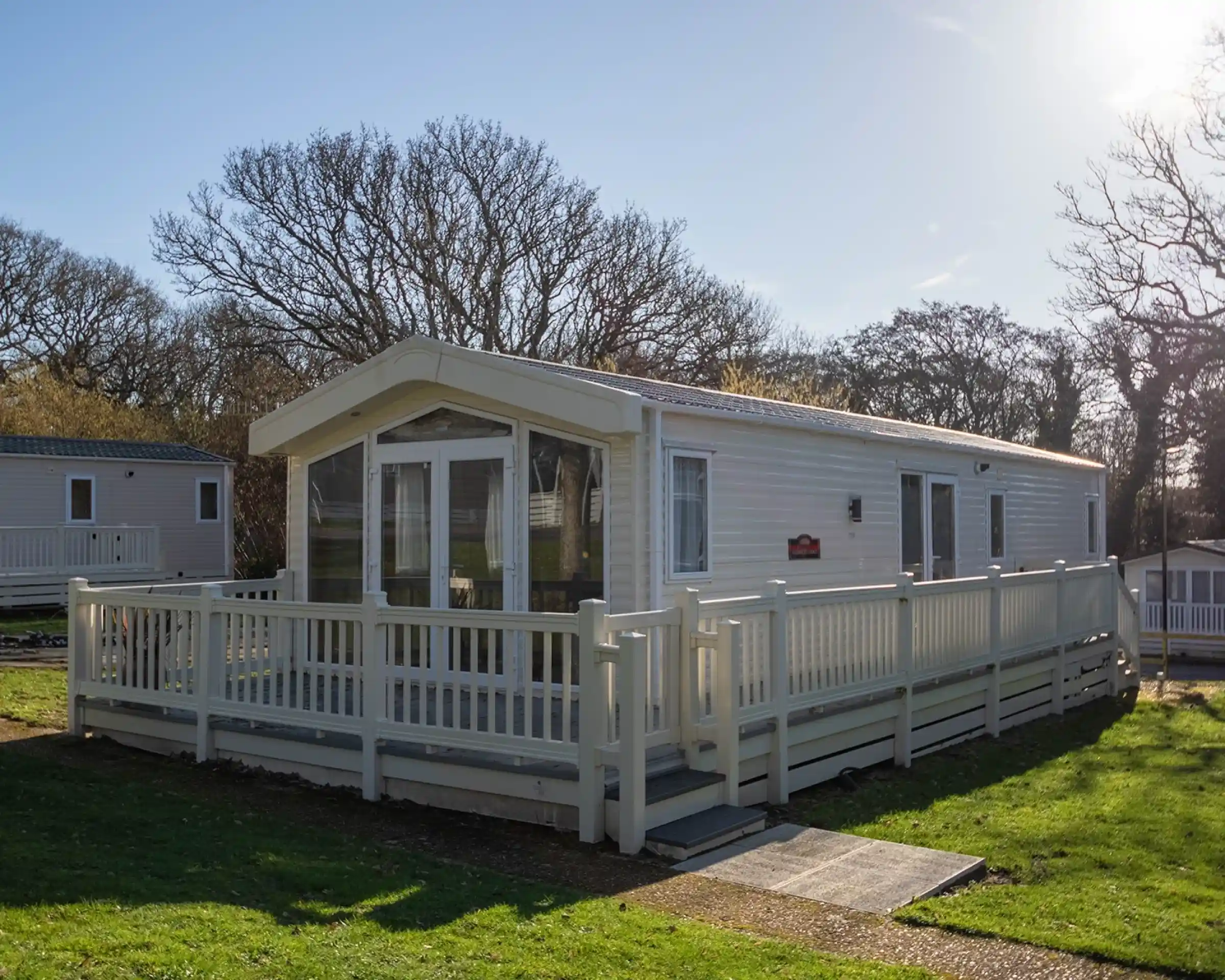 A modern white mobile home with a large deck surrounded by lush green grass and trees, set against a clear blue sky.