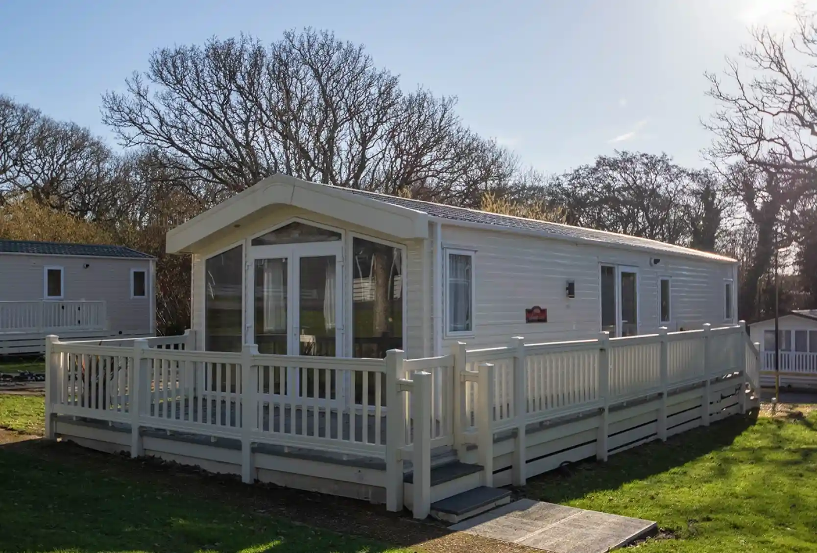 A modern white mobile home with a large deck surrounded by lush green grass and trees, set against a clear blue sky.