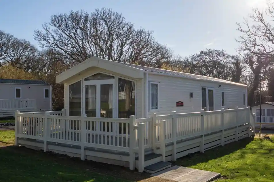 A modern white mobile home with a large deck surrounded by lush green grass and trees, set against a clear blue sky.