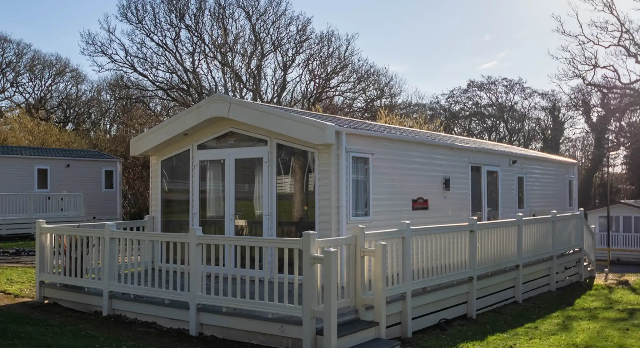 A modern white mobile home with a large deck surrounded by lush green grass and trees, set against a clear blue sky.
