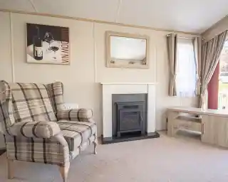 A cozy living room featuring a plaid armchair, a modern electric fireplace with a sleek mantle, and a light wood coffee table. A large window lets in natural light, and a wine-themed artwork is displayed on the wall.