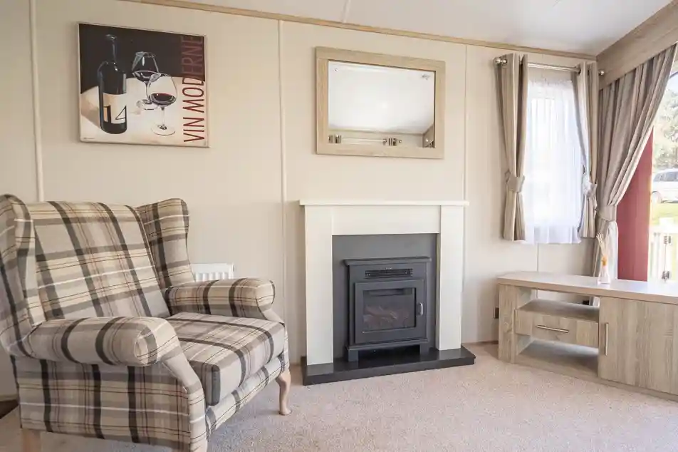 A cozy living room featuring a plaid armchair, a modern electric fireplace with a sleek mantle, and a light wood coffee table. A large window lets in natural light, and a wine-themed artwork is displayed on the wall.