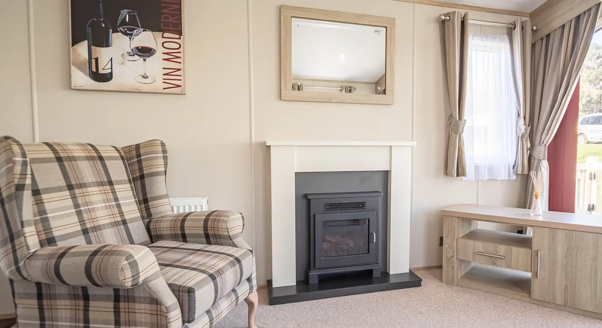 A cozy living room featuring a plaid armchair, a modern electric fireplace with a sleek mantle, and a light wood coffee table. A large window lets in natural light, and a wine-themed artwork is displayed on the wall.