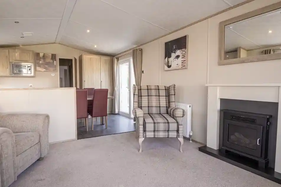 A cozy living room featuring a plaid upholstered armchair, a small fireplace, and a mirror on the wall. The space includes a light carpet and a dining area in the background with red upholstered chairs.