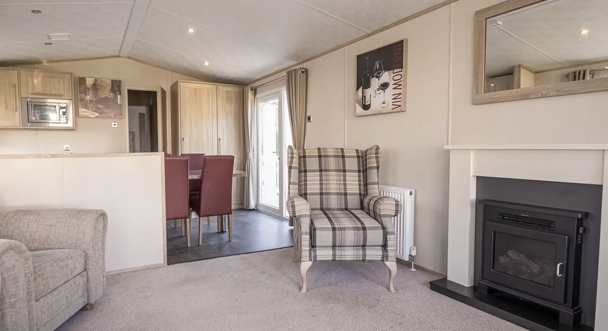 A cozy living room featuring a plaid upholstered armchair, a small fireplace, and a mirror on the wall. The space includes a light carpet and a dining area in the background with red upholstered chairs.