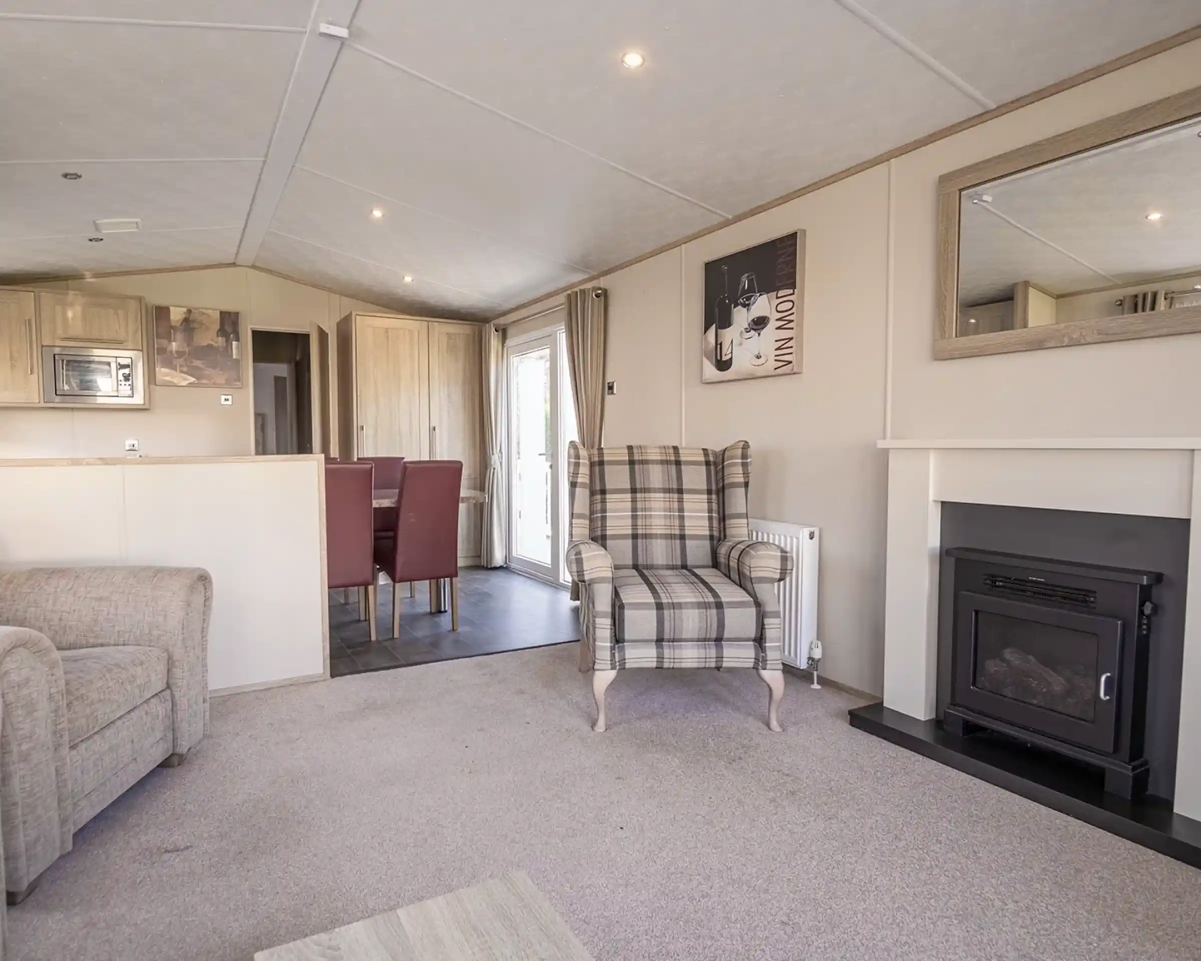 A cozy living room featuring a plaid upholstered armchair, a small fireplace, and a mirror on the wall. The space includes a light carpet and a dining area in the background with red upholstered chairs.
