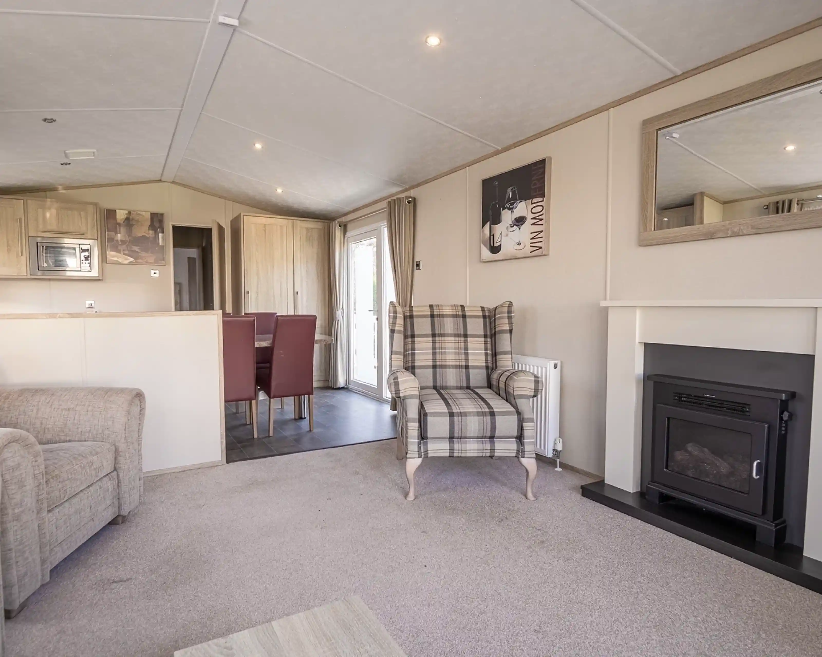 A cozy living room featuring a plaid upholstered armchair, a small fireplace, and a mirror on the wall. The space includes a light carpet and a dining area in the background with red upholstered chairs.
