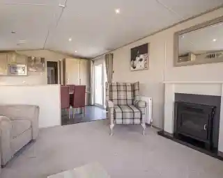A cozy living room featuring a plaid upholstered armchair, a small fireplace, and a mirror on the wall. The space includes a light carpet and a dining area in the background with red upholstered chairs.