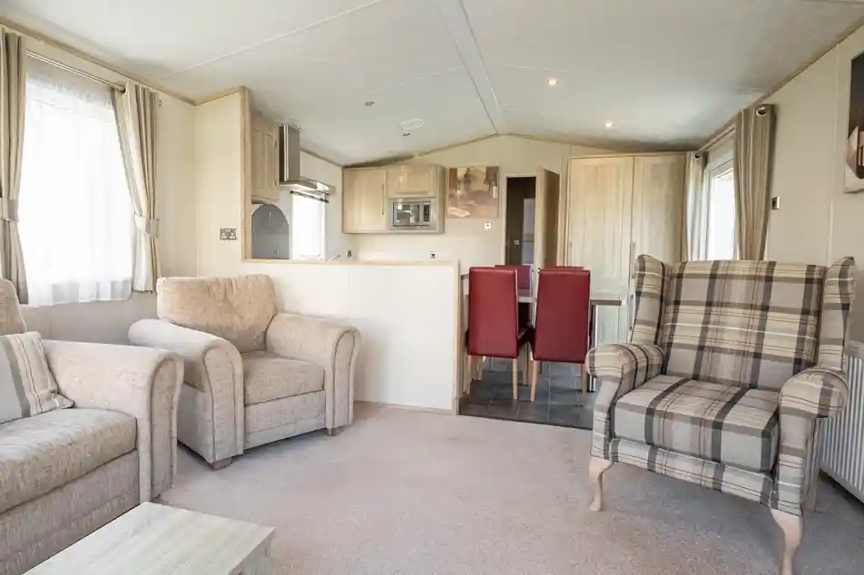 A cozy living area featuring two beige armchairs, a plaid patterned armchair, and a small coffee table. In the background, there’s a dining area with a table and red chairs, along with a modern kitchen equipped with light wood cabinets. Soft natural light filters through the curtains.