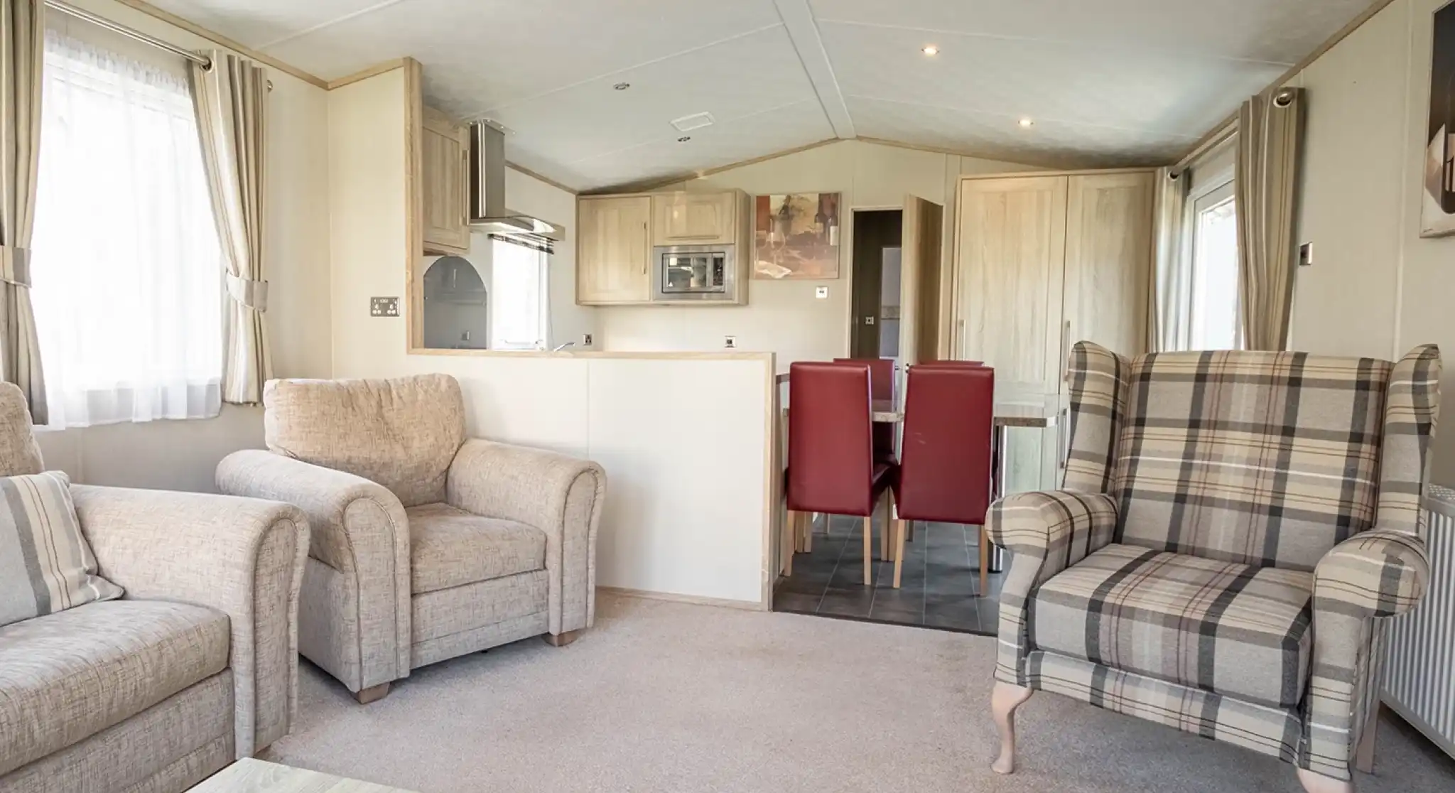 A cozy living area featuring two beige armchairs, a plaid patterned armchair, and a small coffee table. In the background, there’s a dining area with a table and red chairs, along with a modern kitchen equipped with light wood cabinets. Soft natural light filters through the curtains.