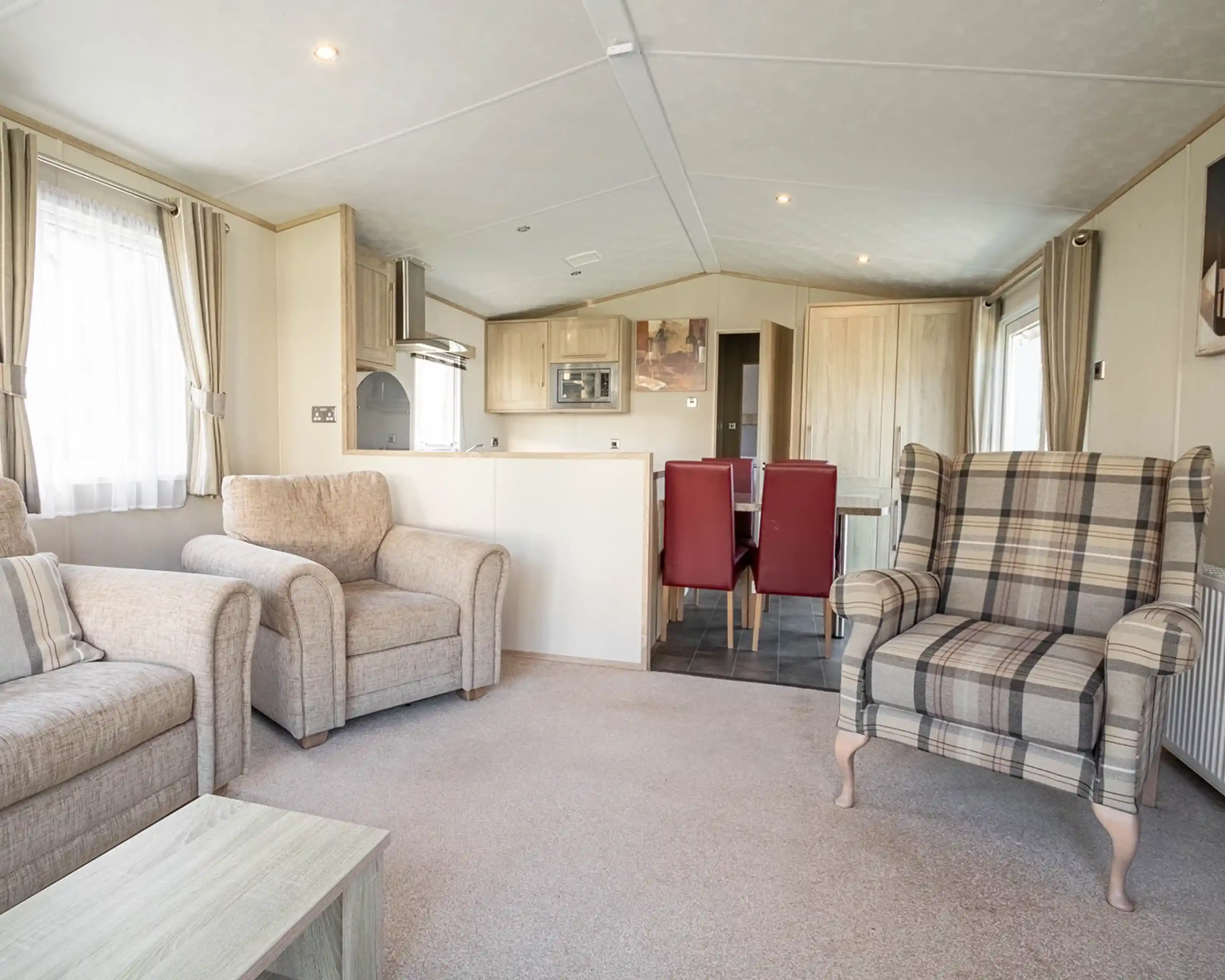 A cozy living area featuring two beige armchairs, a plaid patterned armchair, and a small coffee table. In the background, there’s a dining area with a table and red chairs, along with a modern kitchen equipped with light wood cabinets. Soft natural light filters through the curtains.