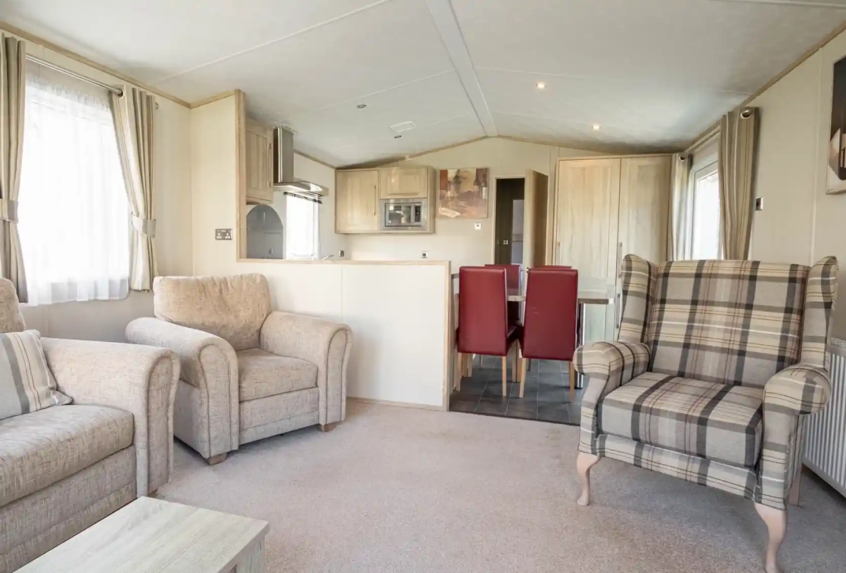 A cozy living area featuring two beige armchairs, a plaid patterned armchair, and a small coffee table. In the background, there’s a dining area with a table and red chairs, along with a modern kitchen equipped with light wood cabinets. Soft natural light filters through the curtains.