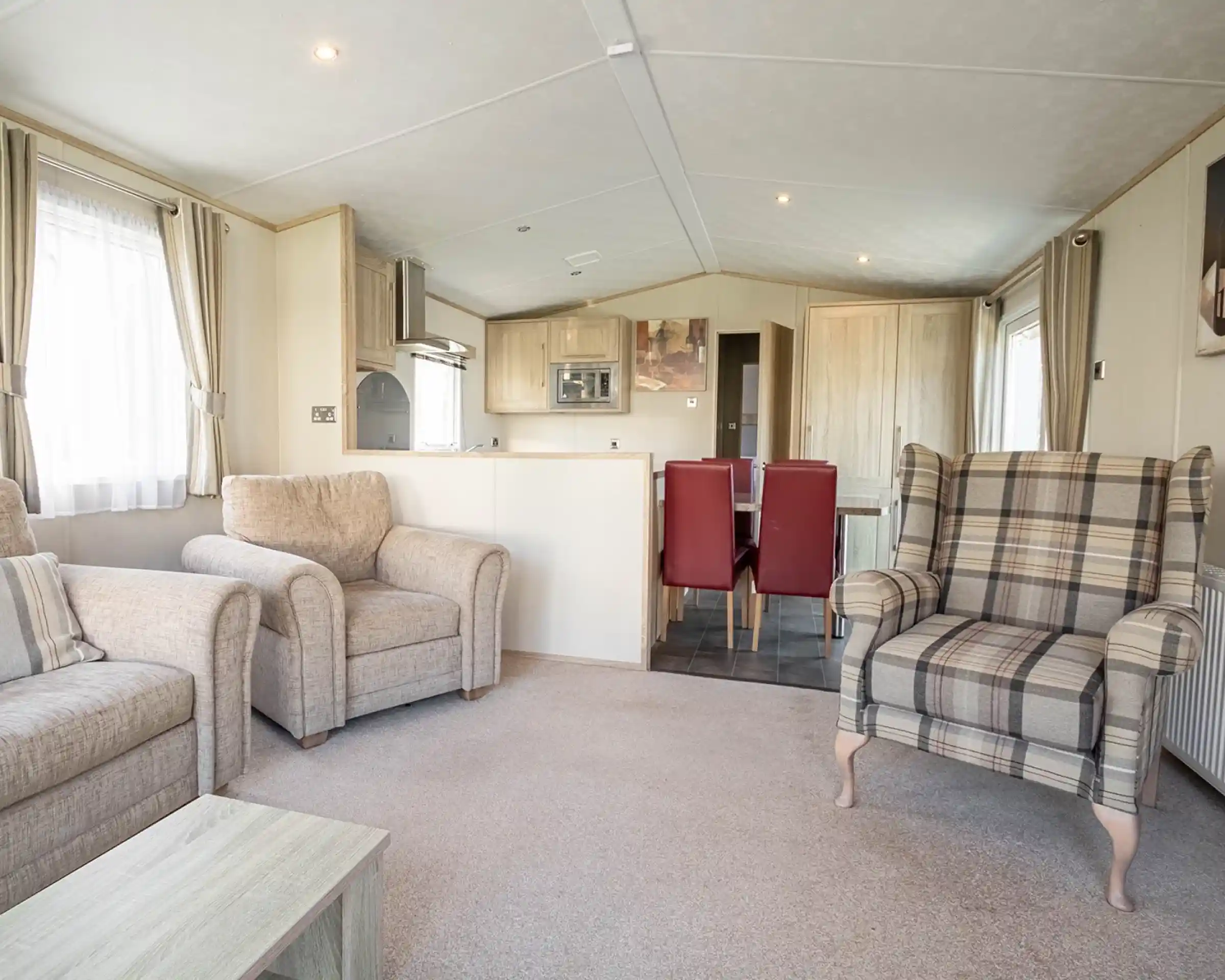 A cozy living area featuring two beige armchairs, a plaid patterned armchair, and a small coffee table. In the background, there’s a dining area with a table and red chairs, along with a modern kitchen equipped with light wood cabinets. Soft natural light filters through the curtains.