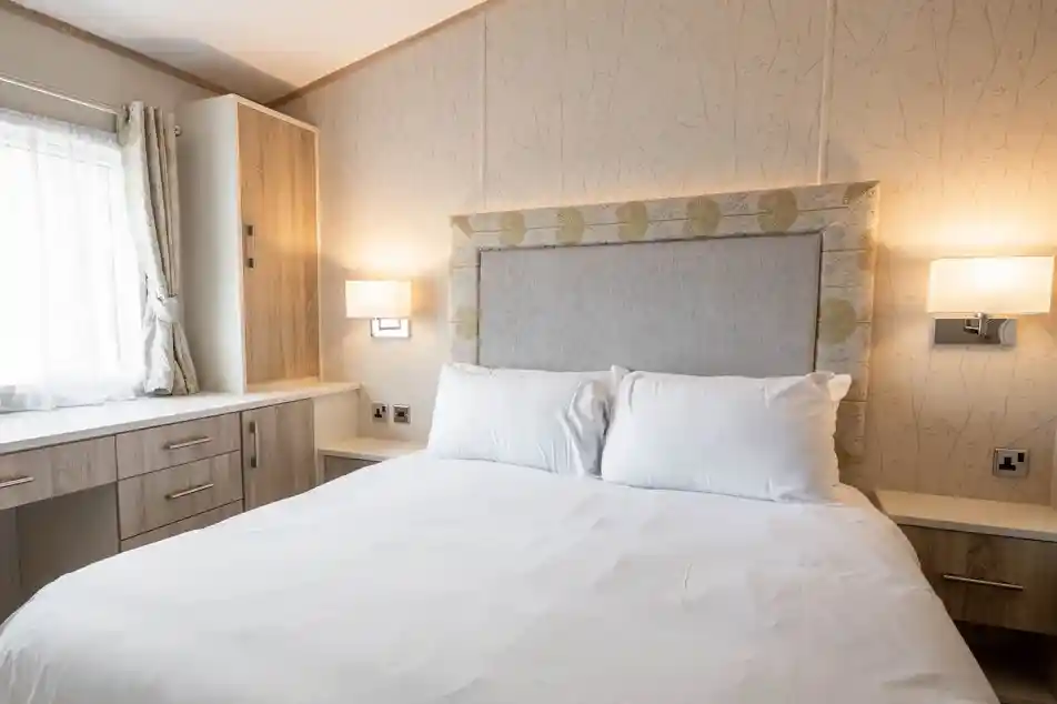 A cozy bedroom featuring a queen-sized bed with white linens, a decorative headboard, and two wall-mounted lamps. A window with light curtains and a wooden dresser or cabinet is visible in the background.