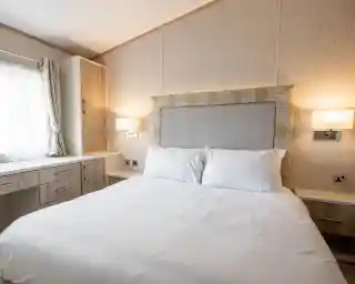 A cozy bedroom featuring a queen-sized bed with white linens, a decorative headboard, and two wall-mounted lamps. A window with light curtains and a wooden dresser or cabinet is visible in the background.