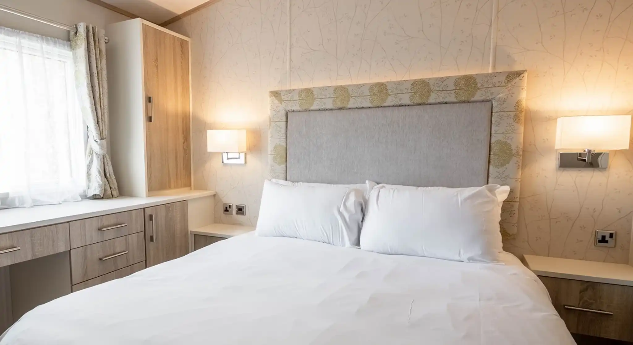 A cozy bedroom featuring a queen-sized bed with white linens, a decorative headboard, and two wall-mounted lamps. A window with light curtains and a wooden dresser or cabinet is visible in the background.