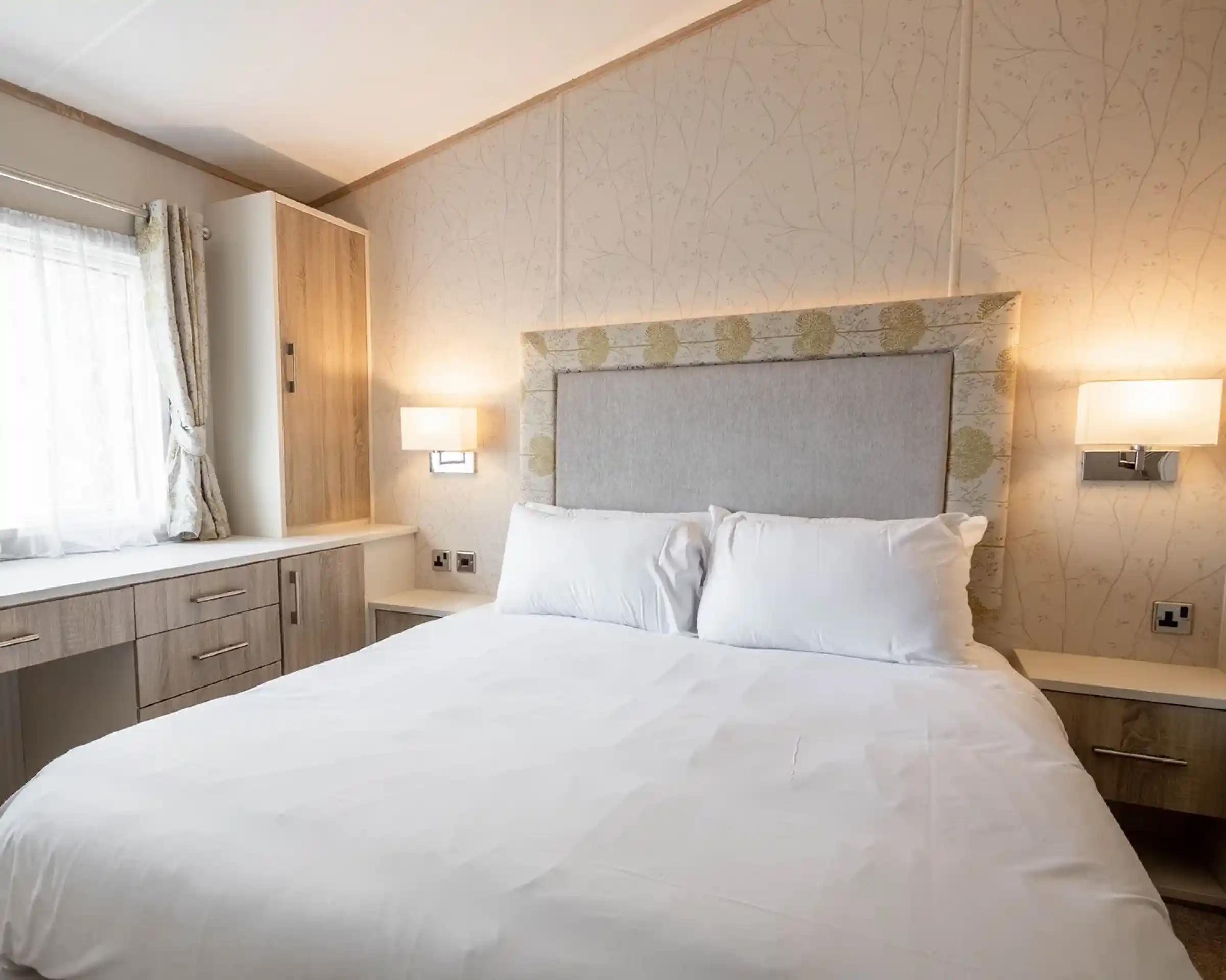 A cozy bedroom featuring a queen-sized bed with white linens, a decorative headboard, and two wall-mounted lamps. A window with light curtains and a wooden dresser or cabinet is visible in the background.