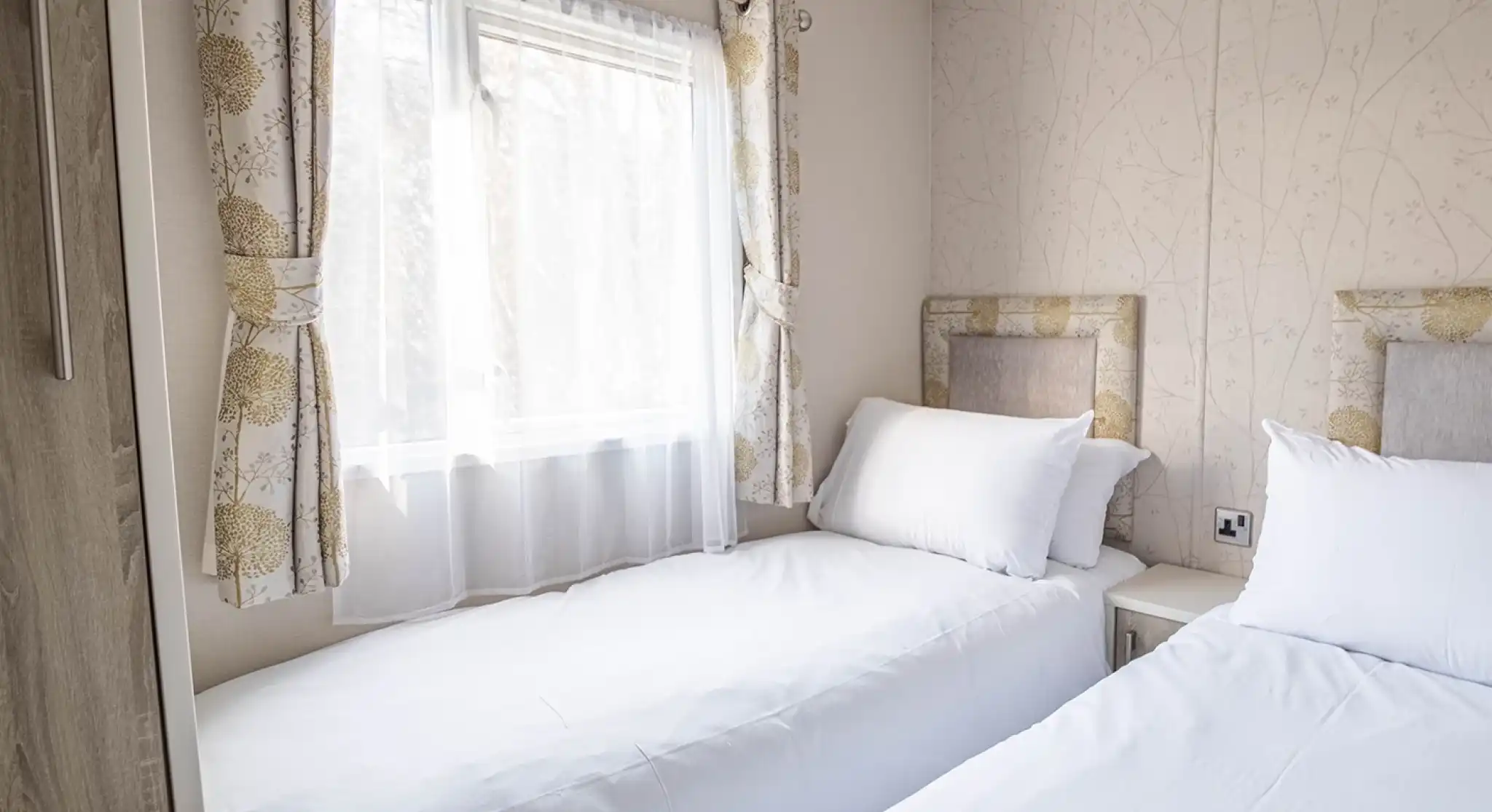 A cozy bedroom with two twin beds topped with white linens and pillows. Natural light streams through a window adorned with sheer curtains, and floral-patterned wallpaper adds a touch of elegance to the space. A nightstand is positioned between the beds.