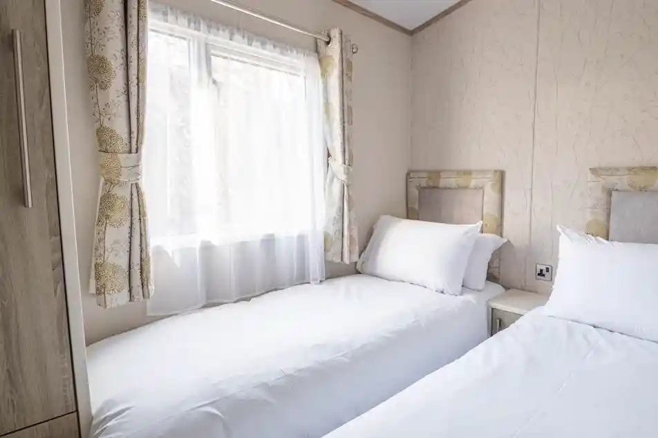 A cozy bedroom with two twin beds topped with white linens and pillows. Natural light streams through a window adorned with sheer curtains, and floral-patterned wallpaper adds a touch of elegance to the space. A nightstand is positioned between the beds.