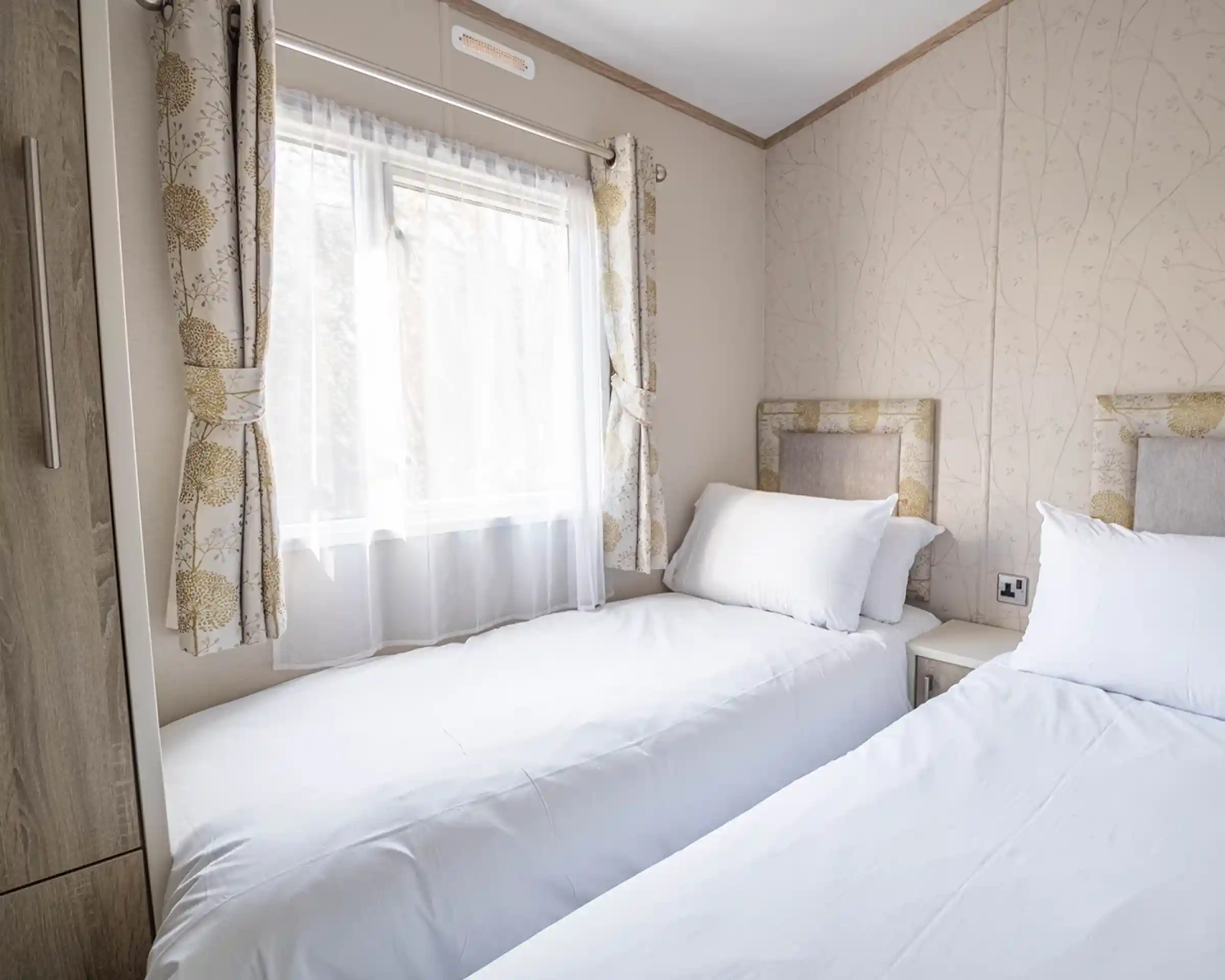 A cozy bedroom with two twin beds topped with white linens and pillows. Natural light streams through a window adorned with sheer curtains, and floral-patterned wallpaper adds a touch of elegance to the space. A nightstand is positioned between the beds.