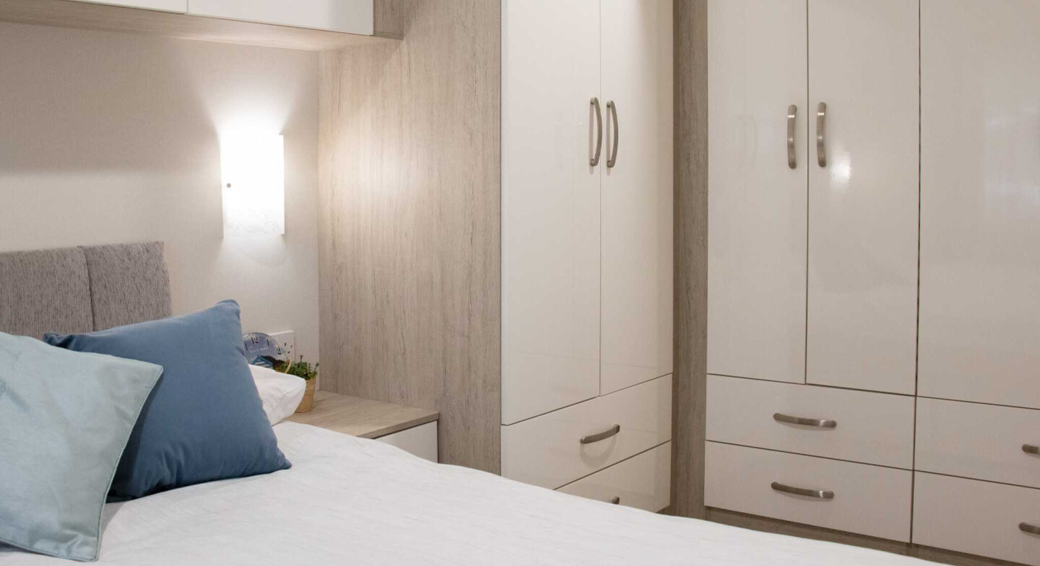 A cozy bedroom featuring a neatly made bed with light bedding and decorative pillows. There is a modern wardrobe with glossy white doors, a bedside table, and soft lighting illuminating the space, creating a welcoming atmosphere.