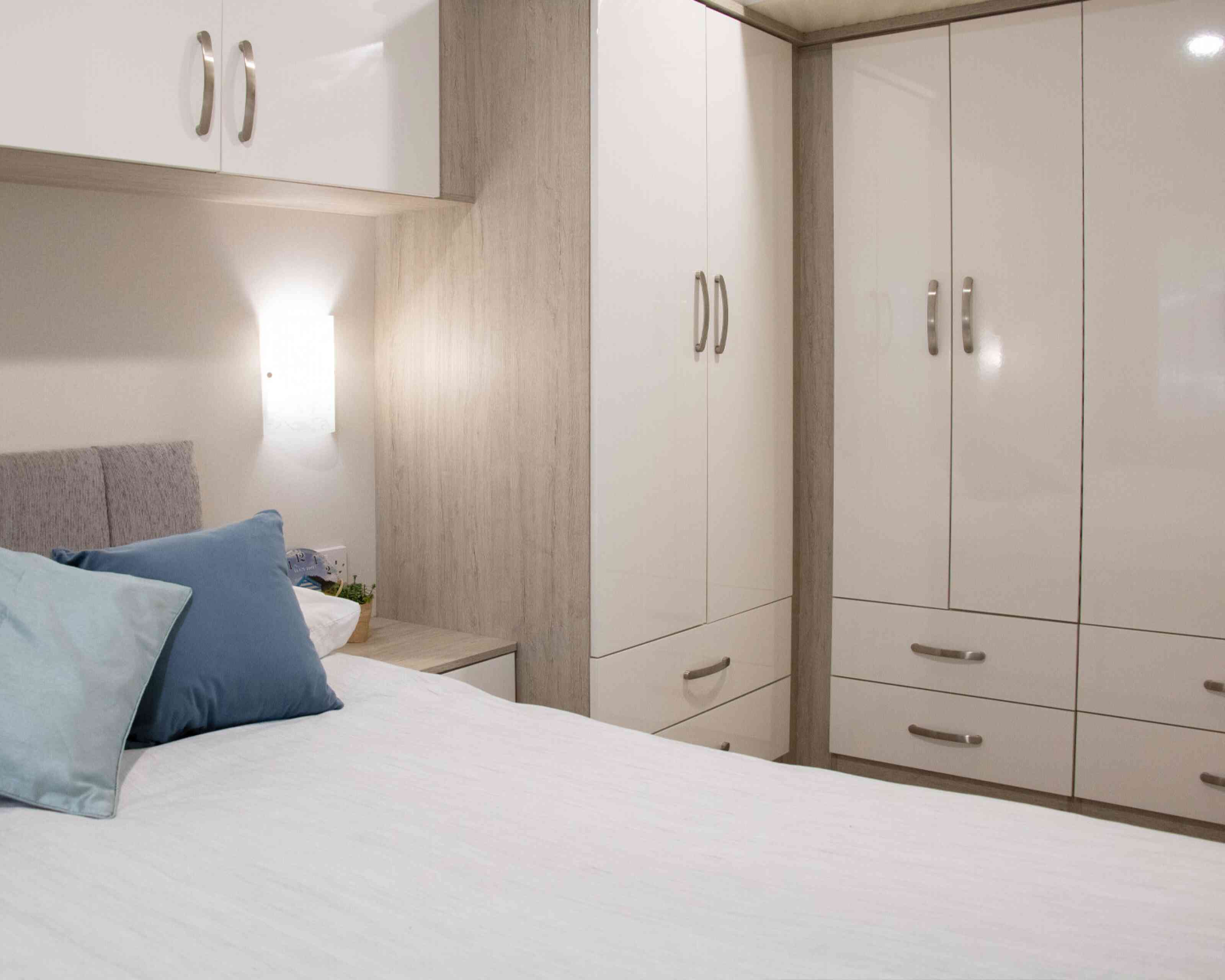 A cozy bedroom featuring a neatly made bed with light bedding and decorative pillows. There is a modern wardrobe with glossy white doors, a bedside table, and soft lighting illuminating the space, creating a welcoming atmosphere.